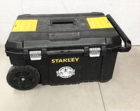 Black Stanley wheeled mobile tool chest with telescoping handle and yellow latch covers, closed, front-side view.