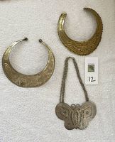 Photo displaying two metal bib necklaces laid flat, one silver-tone with floral and linear engravings, the other gold-tone with geometric details, alongside a butterfly-shaped metal statement necklace with ornate cut-out patterns and attached chain.