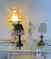 Photo of four electric lamps on glass table showing a tall oil lamp style light, small Tiffany-style stained glass lamp, clear cut glass lamp, and brass lamp with beige shade
