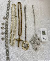 Five necklaces displayed on a white towel, showing varying designs and metals including flower chain, faux pearl, gold cross, mesh-style pendant, and silver-tone dangle pendant.