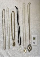 Photo of six necklaces arranged vertically on a light background including two silver tone chains, one gold tassel pendant on chain, one crystal pendant on black cord, one silver teardrop pendant on chain, and one red rhinestone silver pendant on chain.
