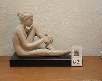 Ceramic ballerina sculpture seated, adjusting pointe shoe, on black base.