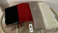 Three folded shawls displayed on a glass table showing colors black/red, beige/white, and solid off-white with fringed edges.