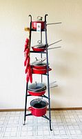 Full view of the black metal pot rack holding the nine-piece red Chantal cookware with glass lids and the 27-inch pottery chili pepper strand hanging on the side.