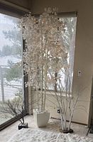 Photo showing three artificial trees near windows: tallest white leafy tree with working white Christmas lights, medium white bare-branch tree with non-working lights, and small black bare-branch tree