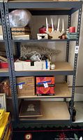 Photo of one metal shelving unit showing four particle board shelves holding various items such as glassware, candles, boxes, and decor items for condition and structure assessment.