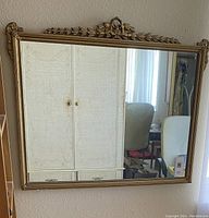 Front view of rectangular mirror showing detailed decorative gold-tone frame with leaf and flower motifs at top center. The mirror glass reflects a room.