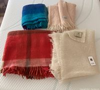 Top-down view of four folded blankets on white surface: blue-green plaid, light peach, red plaid, and off-white.