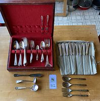 Flatware pieces arranged on table showing Louis XVI pattern set in wood case with red velvet interior, Morningstar and Adam pattern pieces rolled in cloth wrap and loose pieces on table.