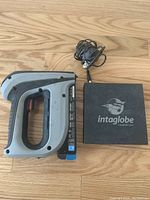 Arrow T50 cordless staple gun next to black charger with cable on wood floor.