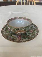 Side and top view of Chinese Famille Rose Canton teacup on saucer showing intricate hand-painted floral and decorative patterns in pink, green, and gold colors.