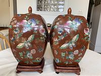 Front view of the two identical brown glazed porcelain jars with butterfly and flower decorations on wooden stands.