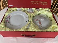 Porcelain tableware set arranged in red gift box with yellow satin lining, showing large plates, covered dish, smaller plates and bowls with hand-painted peach blossom design.