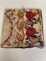Folded silk scarf in original plastic packaging, showing floral and ornate scroll design on cream background with red flowers.