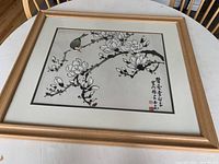 Framed painting showing magnolia blossoms and a bird perched on a branch, with Chinese calligraphy and red seals, laid on a table with visible wood framed edges.