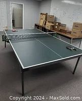 Full view of used full-sized ping pong table with paddles and balls on top, showing net, stand, and surrounding room.