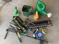 Photo shows various gardening tools laid out on a concrete floor, including a green plastic watering can with a rose sprinkler head, Scotts granular spreader, handheld sprayers, and metal hand tools such as cultivators and pruning shears.