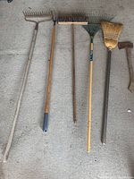 Six garden and yard tools arranged on concrete floor. From left to right: a large metal leaf rake with wooden handle, garden shears with long wooden handles, medium metal tine rake, yellow handled garden fan rake with plastic tines, small broom with natural bristles, and small hatchet.