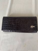 Front view of the dark brown leather clutch wallet showing crocodile embossing and metal Lacoste logo on the flap.