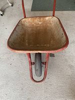Front view of wheelbarrow showing metal beige tub with rust and red frame with single front wheel.