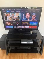 Front view of Sharp 42-inch LCD TV on the metal and glass 2-tier black TV stand, remote on top shelf, TV is on and displaying interface.