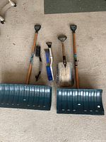Photo showing two large snow shovels with plastic blades and wooden handles, and three small hand snow tools including a blue bristle brush, small scraper brush, and metal shovel.