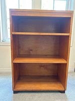 Front view of the small wooden book shelf showing three shelves and decorative trim on top with some visible scratches and wear.