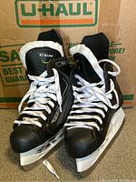 Pair of black CCM hockey skates with white laces and white plastic blade holders visible front and top angles.