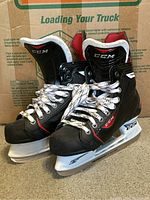 Black CCM hockey skates showing front and side profile with white laces and red-white interior lining, standing on carpet.