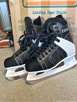 Full side view of black and white CCM hockey skates with white blade holders and metal blades, placed on carpeted floor.
