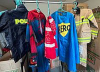Full view of children's costumes hanging on hangers including police, Spiderman, superhero cape, firefighter jacket and pants.