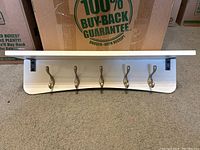 Photo of white wooden wall-mount coat rack with five metal hooks and top shelf, positioned against a carpet floor with boxes behind it.