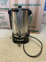 Full view of stainless steel coffee percolator showing the cylindrical body, black handles, hinged lid, black base with power cord and dispensing spigot near the bottom.