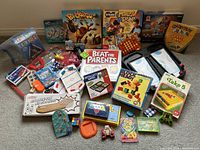 Various children's and family games including board games, card games, puzzles, and small toys, displayed on carpet floor.