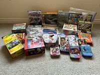 Wide view of all puzzles arranged on carpet, showing the variety of boxed, tin, and case puzzles with varying piece counts from 24 to 1000.