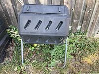 Front view of black dual-compartment rotating compost tumbler on metal legs showing vented panels and lid clips.