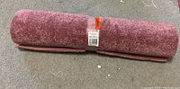 Rolled small dark pink area rug on a gray carpeted floor with clear wrap and orange Loblaws tag showing the price.