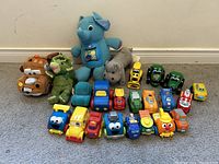 Multiple toddler toys including plush toys and numerous colorful small trucks and character vehicles arranged in rows on carpeted floor