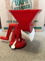 Full view of red Velox tomato press showing hand crank, hopper funnel, suction base and white catch basin