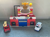 Toy fire station front with fire truck and ambulance beside it on floor