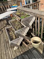 Front view of the wooden tiered herb planter on a deck with plant soil and some remaining dried herbs inside the planting boxes. Nearby empty pots are visible but not included.
