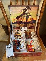 Box open showing four ceramic mugs, each decorated with Tom Thomson painting reproductions, inside a satin-lined commemorative box with a matching print on the inside lid.