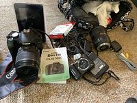 Overview of all lot contents on carpet: Canon DSLR with lens, manual, cables, second lens, strap, filter package, chargers, Samsung camera, bag.