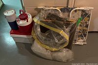 Photo showing two beverage jugs, a cooler box, and a partially packaged foldable lounge chair on basement floor.