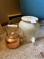 Photo showing ceramic jug with lid and white dispenser spout next to copper kettle on carpeted floor.