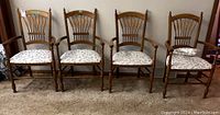 Four medium stained wooden armchairs with carved wheat design backs and upholstered seat pads with matching leaf pattern fabric. One chair has a small matching pillow.