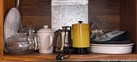 Wide view of shelf with assortment of coffee makers, coffee pots, clear glass bakeware and ceramic pie pans including pie dishes and casserole dishes.