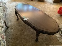 Oval wooden coffee table with smooth top, scalloped edge, and decorative legs on carpeted floor next to floral fabric sofa.