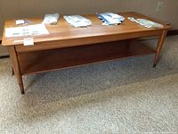 Front angle view of a mid-century modern coffee table with several packets and papers on top, showcasing the rectangular wooden top and lower shelf.