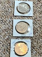 Three Canadian one dollar coins dated 1972, 1982, and 1986 each secured in white cardboard holders on carpeted surface